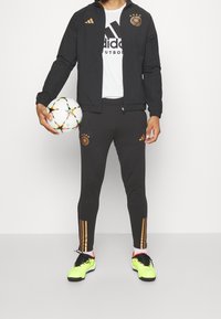 Svart idrottsjacka med guldaccenter, kombinerad med svarta byxor med guldsträck. Modellen håller i en flerfärgad fotboll. Neongröna skor.