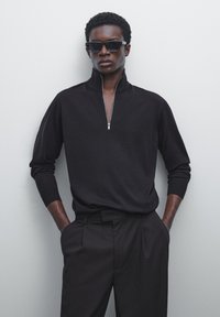 Zwarte gebreide pullover met rits en lange sleeves, gecombineerd met op maat gemaakte zwarte broeken. Soepele textuur en relaxte pasvorm voor een moderne uitstraling.