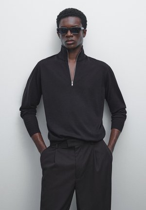Schwarzer, mit Reißverschluss versehener Strickpullover mit langen Ärmeln, kombiniert mit taillierten schwarzen Hosen. Glatte Textur und lässige Passform sorgen für einen modernen Look.