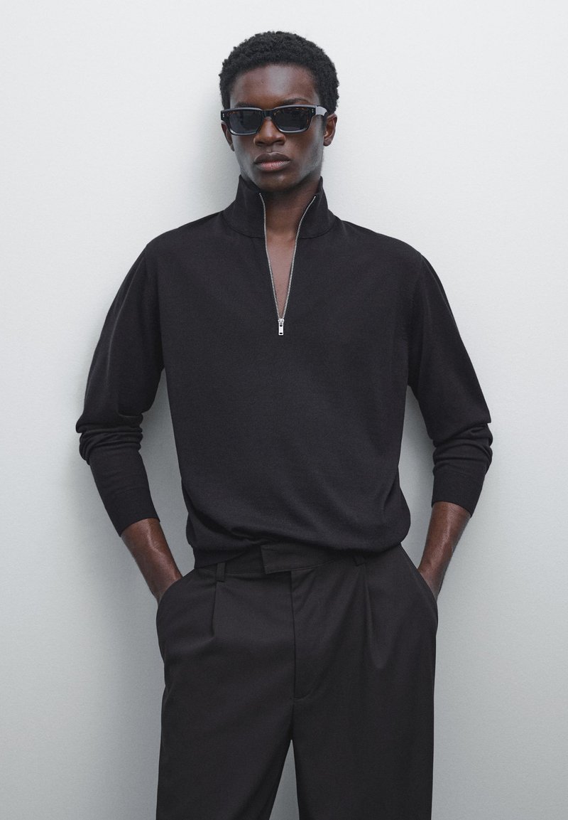Zwarte gebreide pullover met rits en lange sleeves, gecombineerd met op maat gemaakte zwarte broeken. Soepele textuur en relaxte pasvorm voor een moderne uitstraling.