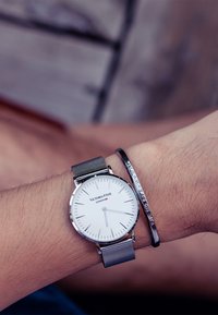 Reloj con correa de malla plateada y una esfera blanca con marcaciones negras, combinado con una elegante pulsera de metal negro. Ambos llevan en una muñeca.
