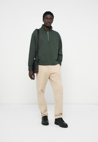Grøn zip-neck pullover, beige bukser og sorte sko. Enkel design med afslappet pasform og subtil tekstur. Sort skuldertaske.