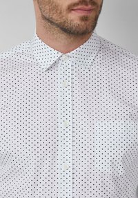 Homme portant une chemise blanche à boutons avec un petit motif à carreaux noirs et une poche sur la poitrine, montrant le bas du visage et une barbe au cou.