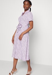 Vestido midi floral em lavanda com mangas curtas, frente com botões, colarinho e cintura ajustada por laço. Tecido suave com uma saia fluida e estampa delicada.