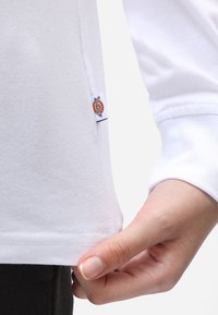 White long-sleeve shirt made of cotton, featuring a straight hem and a small brand tag on the side. The sleeve cuffs are ribbed.