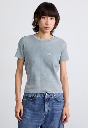 T-shirt en coton ribbé bleu clair avec col rond et manches courtes ; présente un petit logo sur la poitrine. Porté avec un jean bleu.