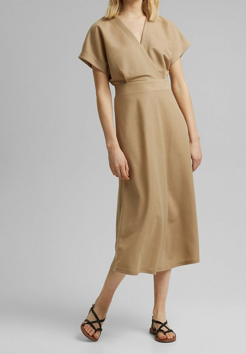 Femme portant une robe midi beige à manches courtes et col en V, associée à des sandales plates noires à brides, debout devant un fond gris uni.