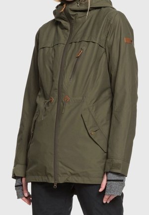 Veste à capuche vert olive avec poche poitrine zippée, taille à cordon, poches latérales à rabat, portée par-dessus des gants gris sans doigts.