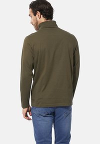 Olivgrünes Langarm-Polohemd mit hohem Kragen, aus weichem Stoff. Über blauen Jeans getragen, zeigt ein einfaches, klares Design.