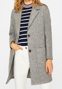 Abrigo de lana gris con solapas de pico, cierre de un solo botón, bolsillos de parche y un corte holgado; combinado con una blusa a rayas en navy y blanco.