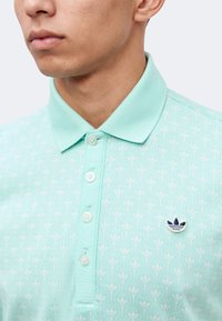 Άνδρας που φοράει ανοιχτό τιρκουάζ πόλο μπλούζα με γιακά με κουμπιά και λευκό floral σχέδιο, με μικρό ναυτικό μπλε και λευκό λογότυπο στο στήθος.