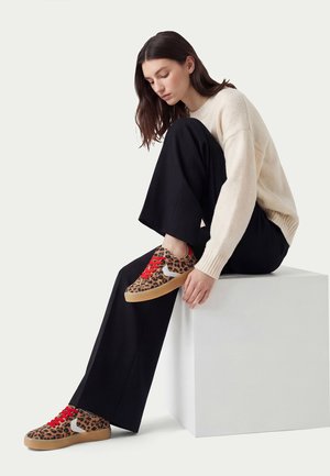 Frau sitzt auf einem weißen Würfel, trägt einen cremefarbenen Pullover, schwarze Hose und Sneakers mit Leopardenmuster und roten Schnürsenkeln, blickt nach unten.