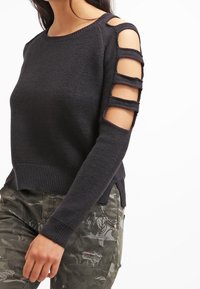 Czarny sweter z dzianiny z otwartymi rękawami, poziomymi wycięciami, prążkowanym dołem oraz długimi, dopasowanymi rękawami. W zestawieniu z spodniami w kamuflażowy wzór.