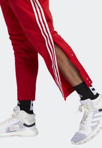 Rode sportbroek met witte zijstrepen en drukknoopsluitingen, gecombineerd met witte en grijze sneakers met gaasaccenten en een zwarte sok.