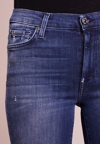 Jeans de mezclilla azul oscuro con cremallera, bolsillo frontal y sutiles detalles desgastados en la tela. Diseño estándar de cinco bolsillos.
