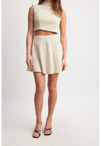 Vaaleanbeige kahden kappaleen asu, jossa on lyhyt, hihaton toppi korkealla kaula-aukolla ja leveä hame. Sileä, teksturoitu kangas.