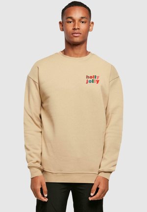 Beige huppari pitkillä hihoilla. Siinä on punainen ja vihreä "holly jolly" -teksti, pyöreä pääntie, resorit hihansuissa ja helmassa. Pehmeä kangas.