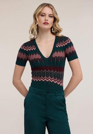 Mujer con top de punto de manga corta y cuello en V profundo con patrón de zigzag en verde, rosa y burdeos, combinado con pantalones verdes oscuros, posando.