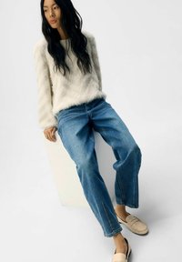 Fjäderlätt gräddvit tröja, ljusblå högmidjade jeans och beige slip-on-skor med svarta accenter. En enkel bakgrund framhäver outfiten.