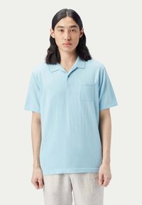 Ljusblå polo-shirt med mjuk textur, korta ärmar, en ficka på vänster bröst och en knappslå med två knappar.