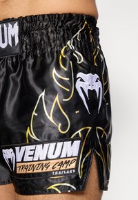 Μαύρα αθλητικά σορτς με κίτρινα και λευκά κυματιστά μοτίβα, ελαστική μέση και λογότυπο Venum Training Camp στο πλάι.