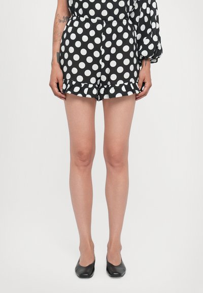 Schwarzer und weißer gepunkteter Jumpsuit mit gerafftem Saum und elastischem Bund. Die Model trägt schwarze Ballerinas.