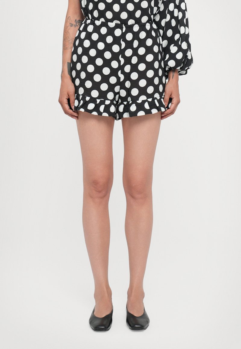 Schwarzer und weißer gepunkteter Jumpsuit mit gerafftem Saum und elastischem Bund. Die Model trägt schwarze Ballerinas.