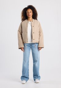 Beige puffer jacket with a button front, two side pockets, and drawstring hem, worn over a white shirt and paired with light blue jeans.