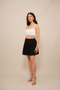 Top corto bianco con pizzo, abbinato a una gonna plissettata nera con un bottone laterale, completato da tacchi con cinturini dorati.