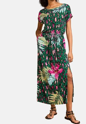 Vrouw die een groene maxi-jurk draagt met roze bloemen- en bladerenprint, korte mouwen, ceintuur in de taille, split aan de zijkant en zwarte sandalen.