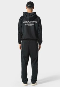 Felpa con cappuccio nera e pantaloni abbinati. Il materiale sembra morbido, con logo e testo bianco sulla schiena. Design semplice, vestibilità casual.