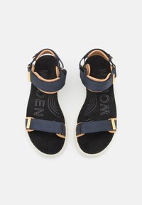 Sandalias de tiras azul marino con correas ajustables en el tobillo y en la parte media del pie, plantillas acolchadas en color marrón, suelas texturizadas en negro y suelas blancas.