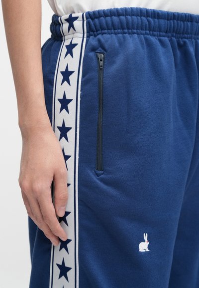 Blue sweatpants made of soft fabric feature a side zip pocket, white side stripes with navy stars, and a small white rabbit logo.