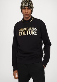 Felpa nera con logo stampato in oro "VERSACE JEANS COUTURE", scollo rotondo e colletto dorato a contrasto. Beanie nero e oro con motivo.