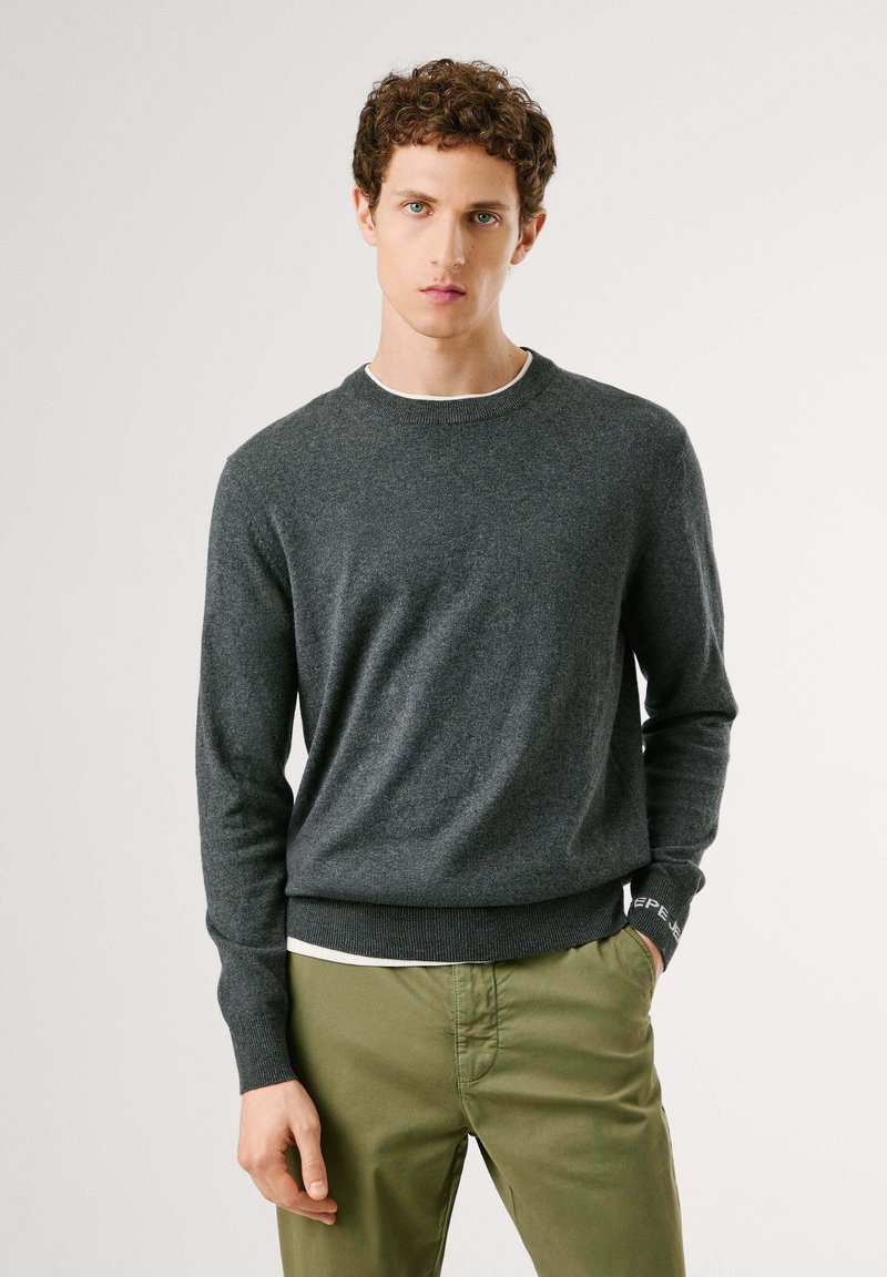 Suéter gris de cuello redondo fabricado con una tela suave, que presenta mangas largas y puños acanalados. Combinado con pantalones de color verde oliva.