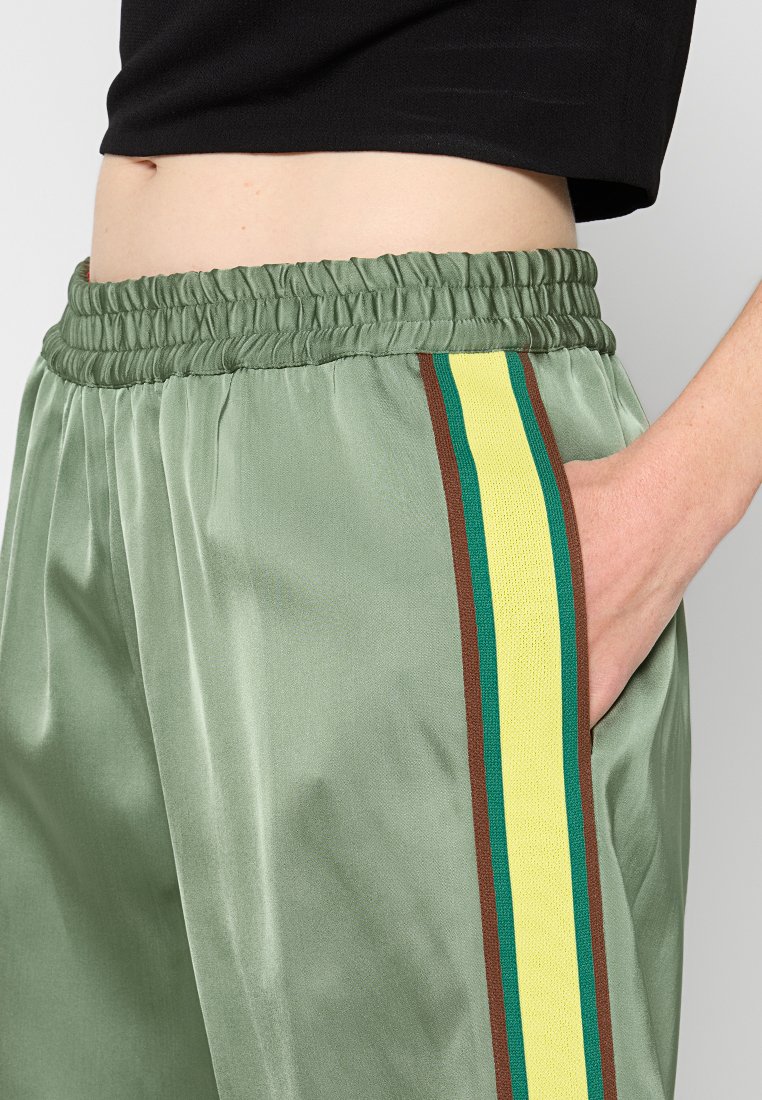 Satin grüne Jogginghose mit seitlichen Taschen, elastischem Bund und dreifachen Streifen in Gelb, Dunkelgrün und Braun an den Seiten.