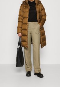 Persoon in lange bruine pufferjas, zwarte shirt, beige broek, zwarte laarzen, houdt met een hand een zwarte tote bag vast terwijl de andere hand in de zak zit.