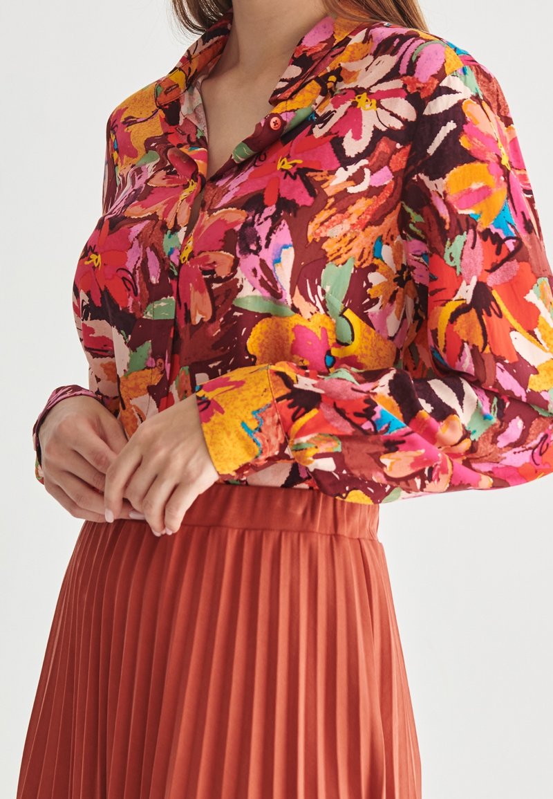 Femme portant un chemisier à manches longues à fleurs vives, rentré dans une jupe plissée taille haute couleur rouille, mains jointes devant elle.