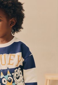 Felpa a righe bianche e blu con personaggi dei cartoni animati e la scritta "BLUEY" in oro. Realizzata in tessuto morbido con scollo rotondo.