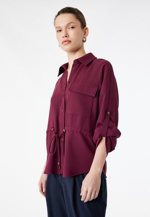 Kobieta o jasnej karnacji i włosach związanych do tyłu, ubrana w bordową koszulę zapinaną na guziki z podwiniętymi rękawami i ściągaczem w talii, zestawioną z ciemnymi spodniami.