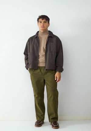 Bruine canvas jacket, beige coltrui, olijfgroene broek en bruine schoenen met decoratieve stiksels. Beschikt over zijzakken en een aansluitend ontwerp.