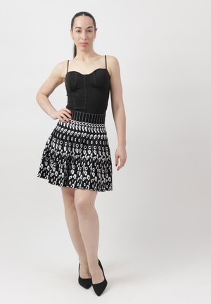 Mujer de pie con la mano en la cadera, vestida con un top negro ajustado y una falda plisada con estampado en blanco y negro, y tacones negros.