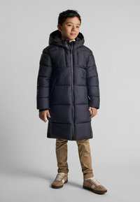 Manteau rembourré noir avec une capuche, présentant des sections matelassées horizontales. Porté avec un pantalon beige et des baskets beiges.