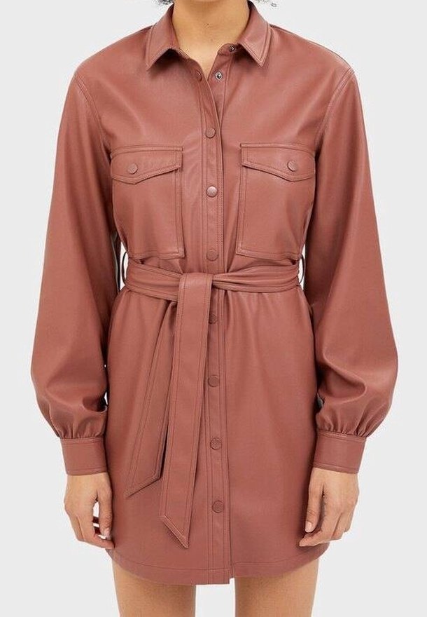 Personne portant une robe-chemise mauve en similicuir, à manches longues, ceinturée et boutonnée, avec deux poches poitrine, debout devant un fond uni.