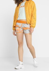 Keltainen verkkobomber-takki, vaaleansininen logollinen t-paita, oranssiraitainen vyötärönauha, harmaa-oranssipainokuvioidut shortsit ja valkoiset urheilukengät.