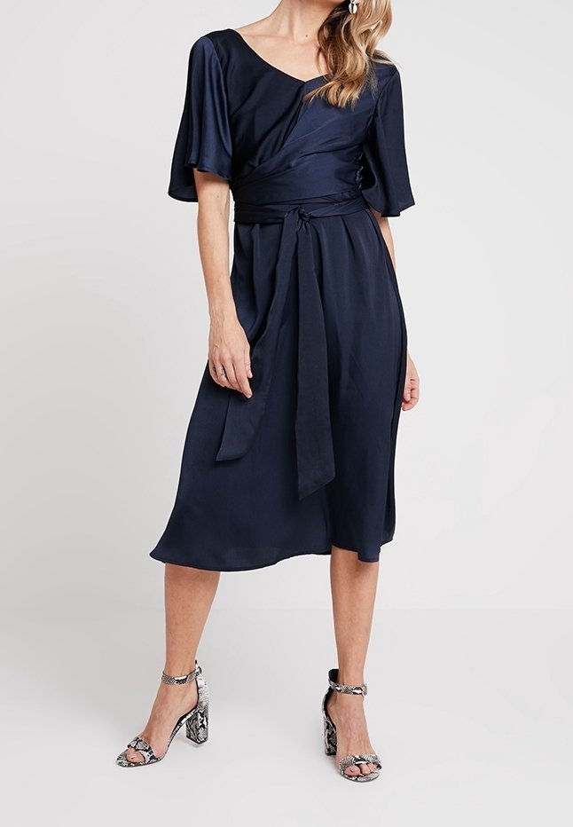 Robe portefeuille marine avec des manches courtes et volantées, taille nouée et longueur mi-mollet. Le mannequin l'associe à des talons argentés à motif serpent.
