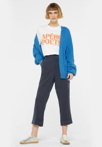 Blauer flauschiger Cardigan über einem weißen T-Shirt mit grafischem Aufdruck in orange, kombiniert mit navyblauen Hosen und metallischen Sneakers.