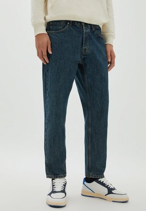 Person wearing dark blue straight-leg jeans, white sneakers with navy accents, and a cream long-sleeve top, standing against a plain background.