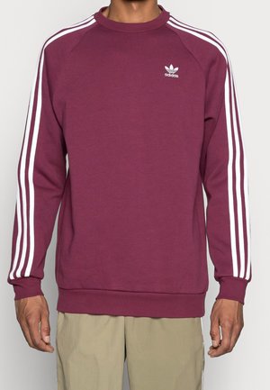 Mężczyzna noszący bordową bluzę Adidas z białymi paskami wzdłuż rękawów i logo Adidas na piersi, w połączeniu z beżowymi spodniami.