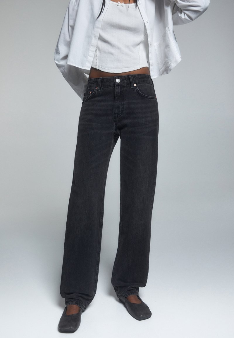 Jeans neri a gamba larga con vita alta, texture sottile e design classico a cinque tasche, abbinati a un top bianco e una camicia bianca ampia.